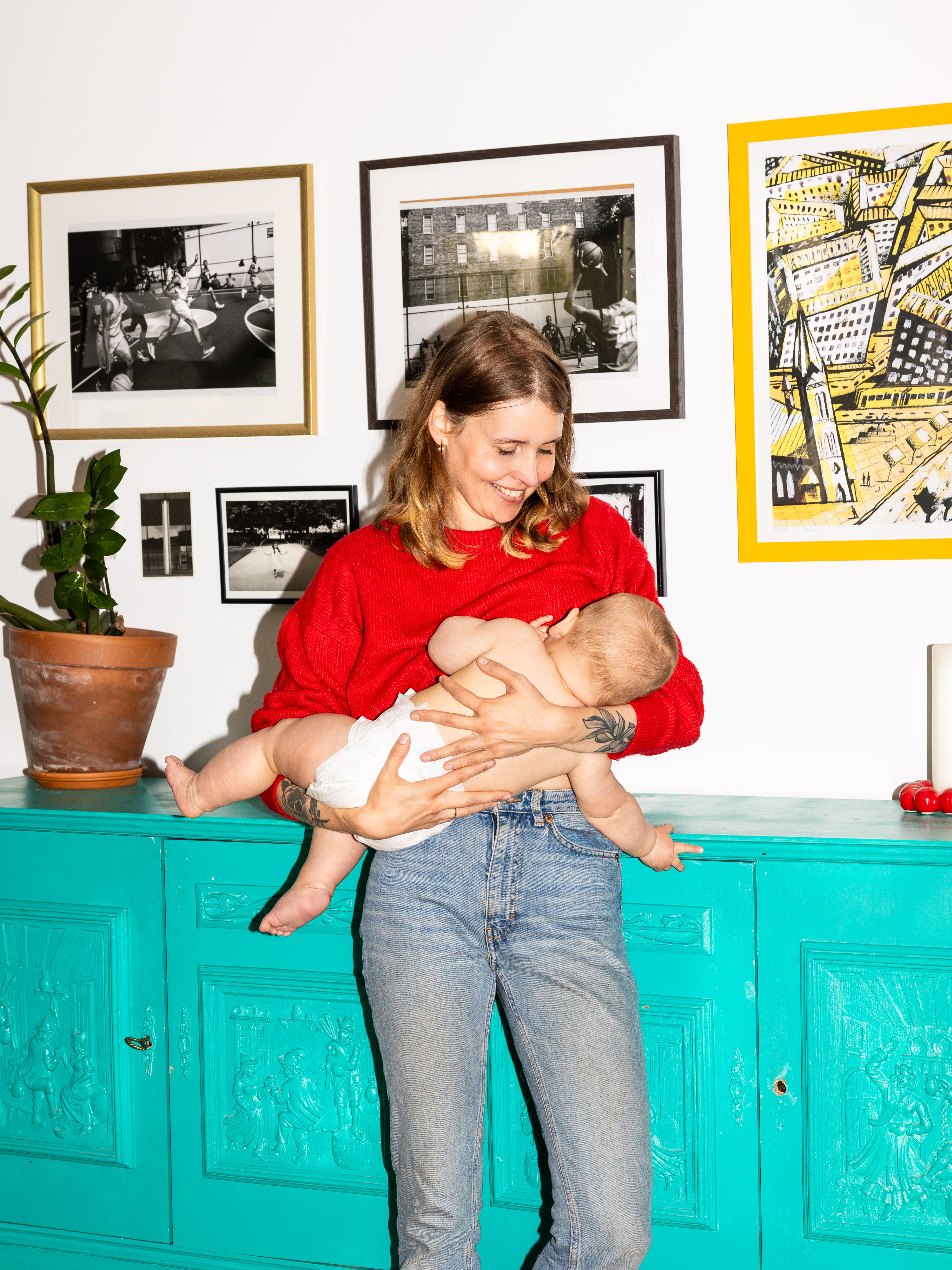 Das Farbfoto zeigt eine junge Frau. Sie hat schulterlange, blonde Haare, trägt einen roten Pullover und Jeans. Sie hält ein Baby im Arm, das sie gerade stillt. Das Baby trägt eine Windel und hat eine entspannte Haltung. Die Frau lächelt liebevoll und schaut das Kind an. Hinter ihr sieht man eine türkisgrüne Kommode. Darüber hängen mehrere gerahmte Schwarz-Weiß-Fotografien und ein Kunstdruck mit einer abstrakten Stadtansicht.