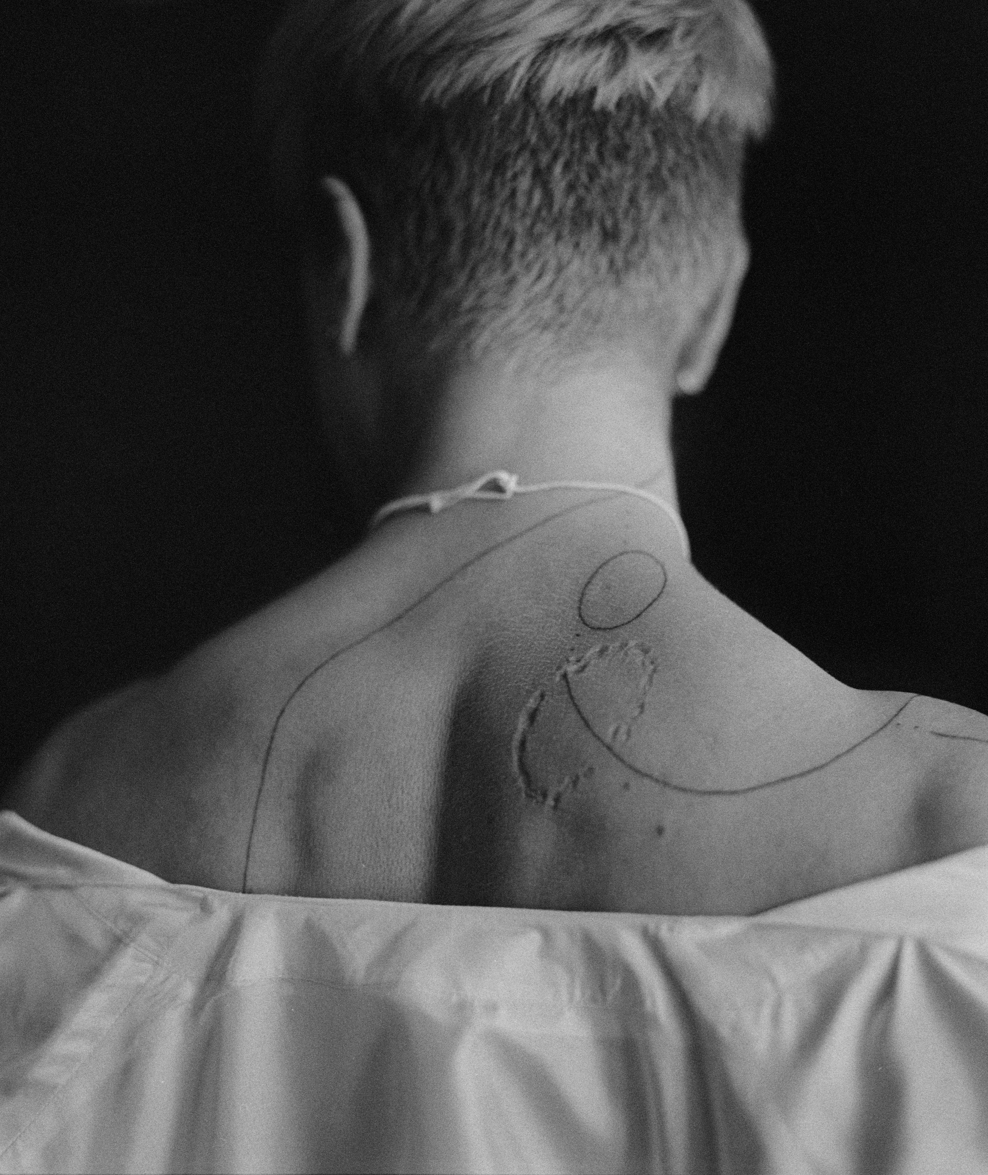 Das Schwarz-Weiß-Foto zeigt Nacken und Rücken einer Person mit kurzem Haar. Die leicht herabgerutschte Bluse gibt die Schulterpartie frei. Auf der Haut sieht man handgezeichnete Tattoos aus drei feinen teilweise gepunkteten Linien und einem Kreis. Auf dem rechten Schulterblatt ist eine narbenähnliche Kreisstruktur zu sehen, die wie eine Bissspur aussieht.
