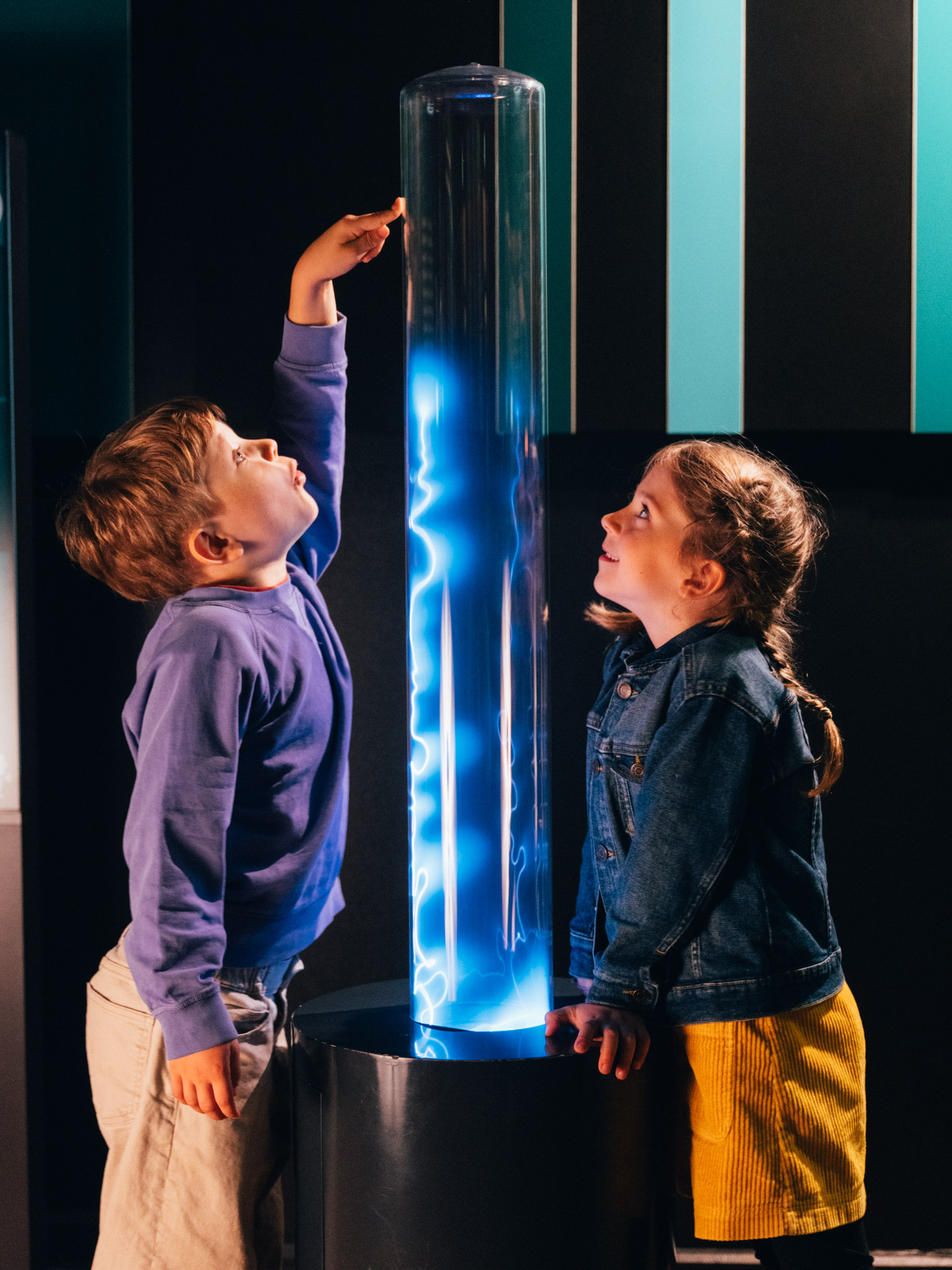 Zwei Kinder stehten in einem dunklen Raum um eine transparente Säule herum. Im Inneren der Säule leuchtet entlang der gesamten Höhe ein blauer Blitz. Eines der Kinder Kinder berührt die Plasmasäule mit seinen Händen.