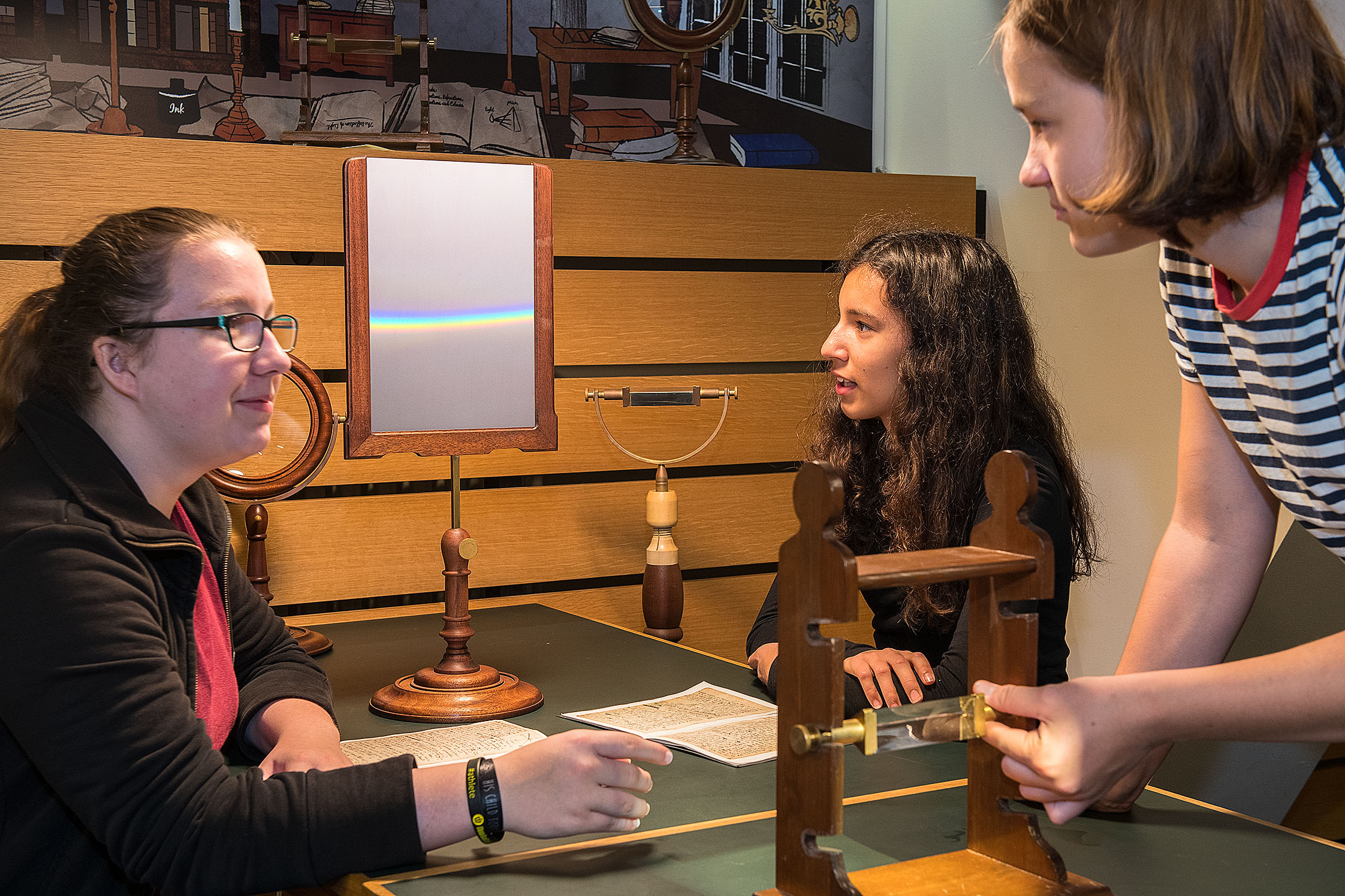 Drei Mädchen experimentieren an einem Tisch. Zwei sitzen sich am Tisch gegenüber, ein drittes steht ganz rechts. Es ist leicht nach vorne gebeugt und justiert ein Glasprisma in einem Holzgestell. Das Mädchen links schaut sie an. Auf dem Tisch steht in einem Holzgestell eine helle Fläche, etwa so groß wie ein A4-Blatt. Auf der Fläche leuchten in einem waagerechten Streifen die Spektralfarben. Das Mädchen in der Mitte betrachtet diese. Die Geräte erwecken durch ihr dunkles Holz und ihre Formen einen historischen Eindruck.