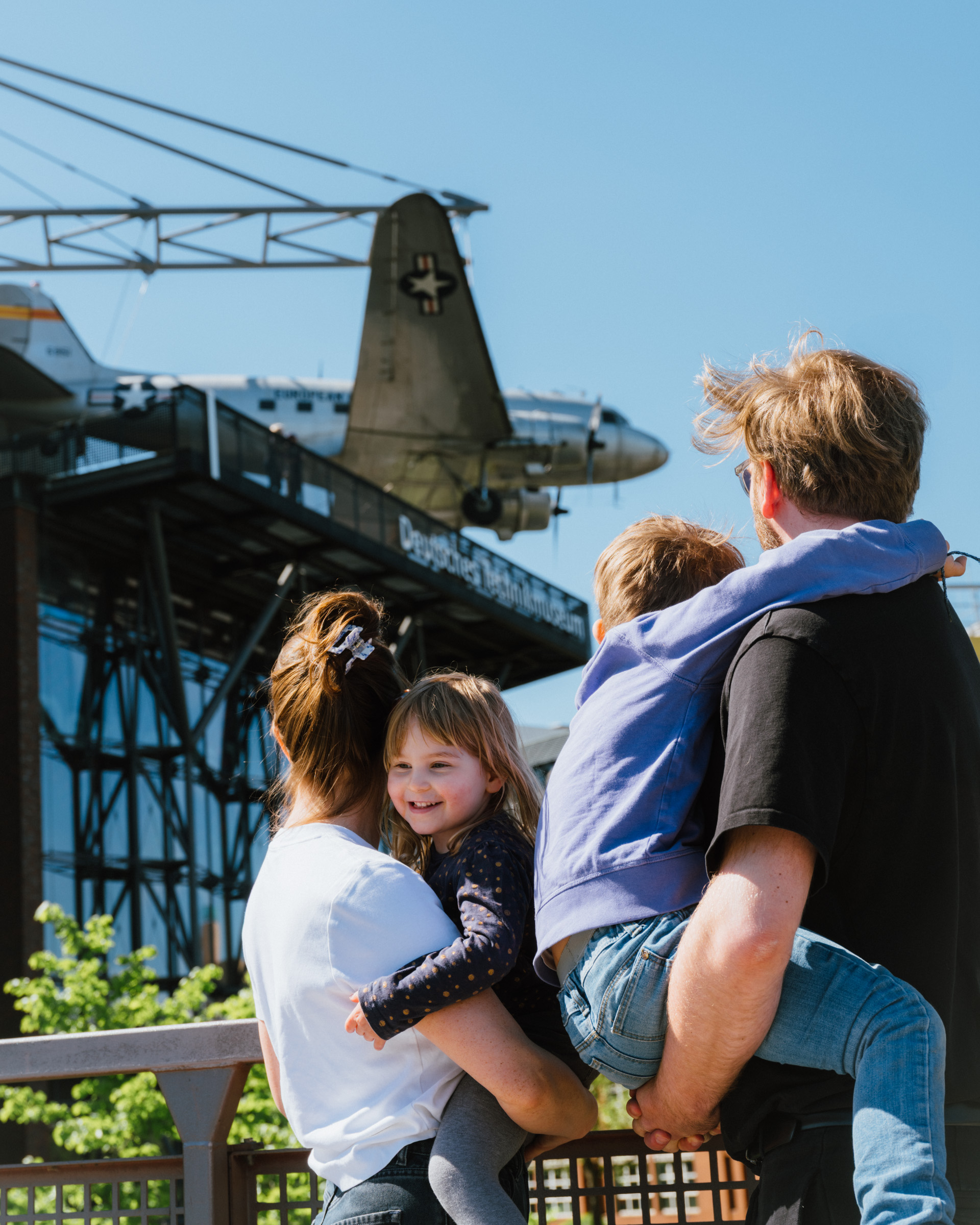 Eine Paar mit zwei Kindern auf dem Arm stehen auf dem Steg gegenüber vom Museum und betrachten das Flugzeug, das dort an der Fassade hängt.