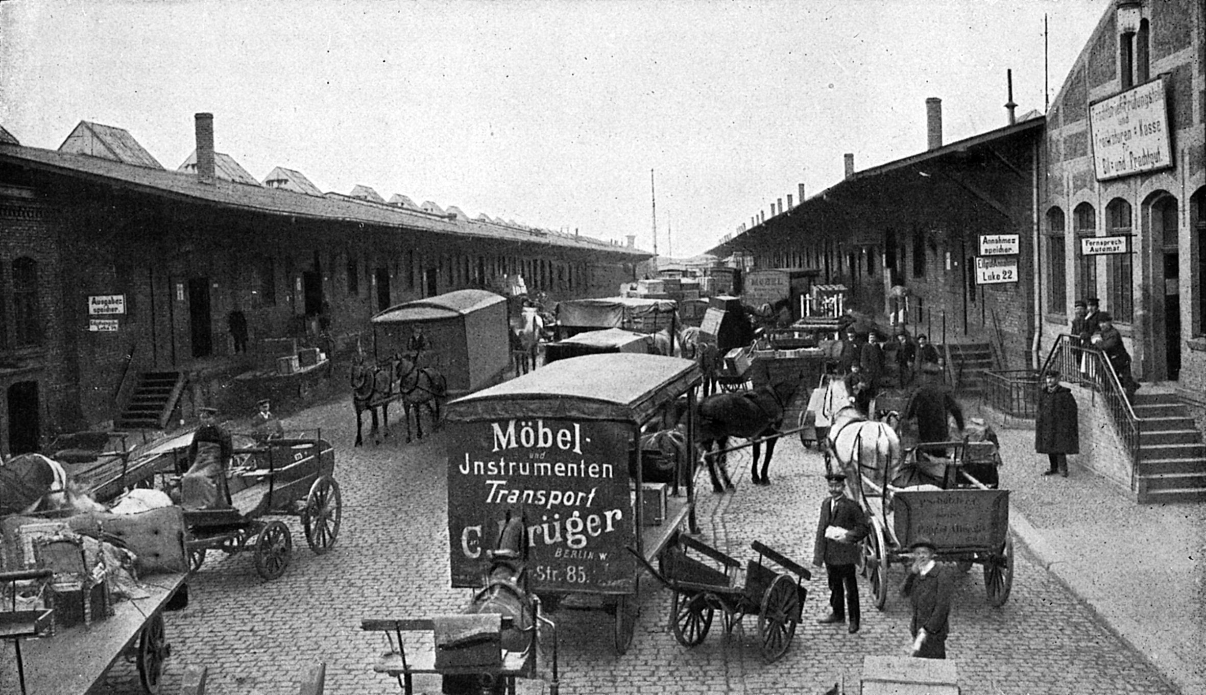 Schwarzweiß-Foto einer kopfsteingepflasterten Straße mit Lagerschuppen auf beiden Seiten. Auf der Straße sind Pferdefuhrwerke, Karren und Menschen unterwegs.