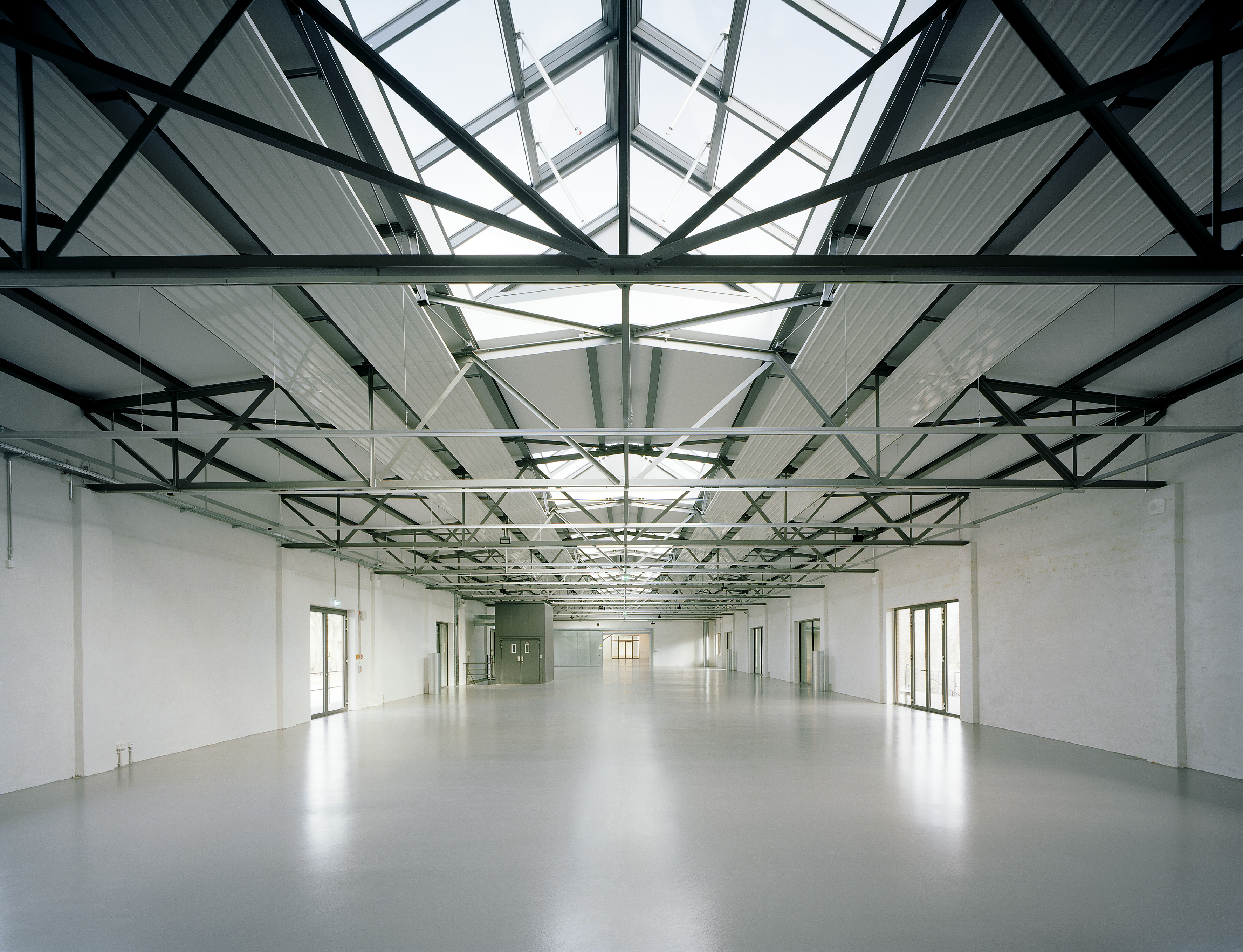 Blick in eine Lagerhalle, die offensichtlich neu restauriert wurde: Die Böden und Wände glänzen weiß, die Stahlkonstruktion der Decken geben den Blick auf Glasgauben frei.