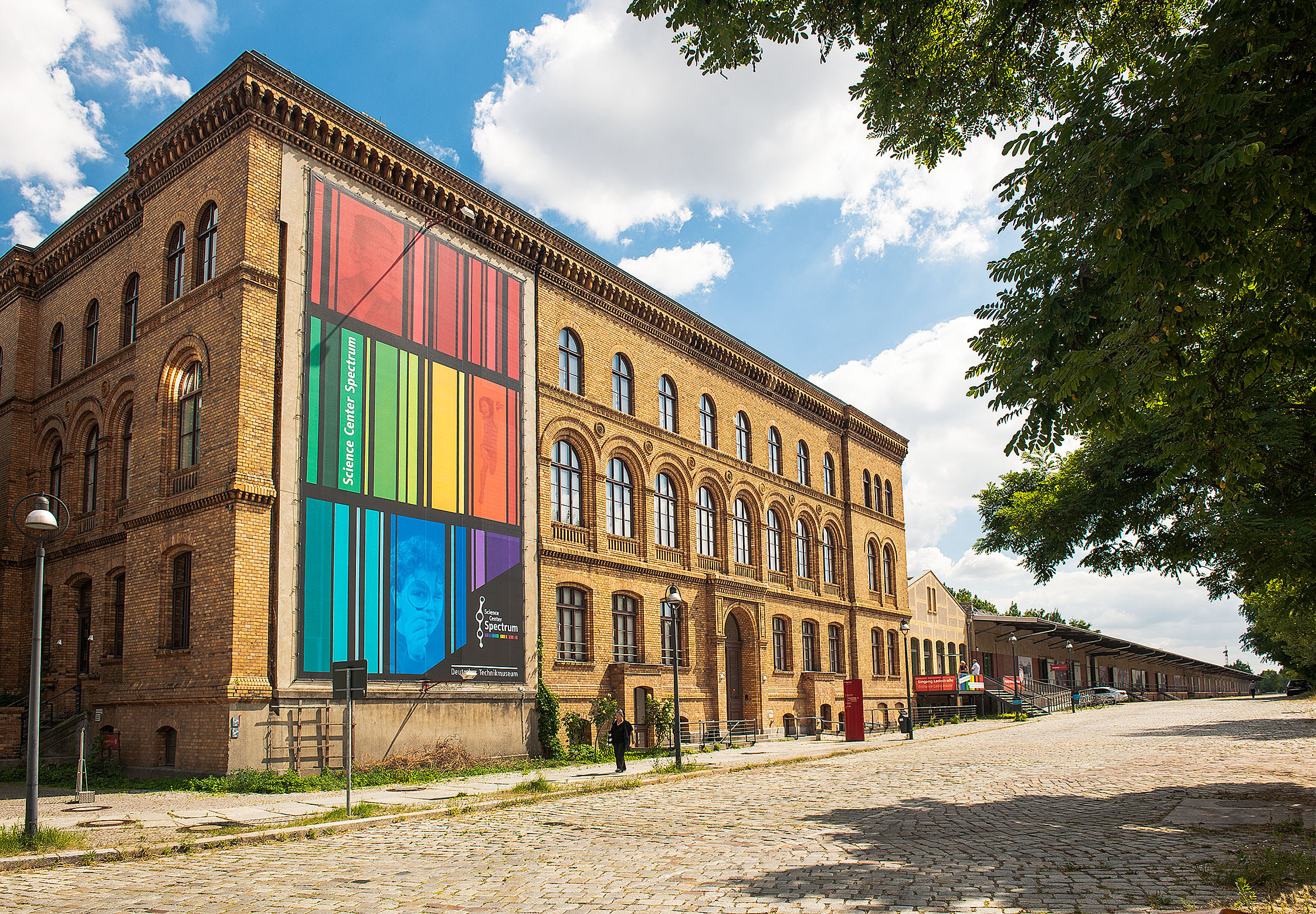 Ein dreigeschossiger Backsteinbau mit einem bunt gestreiften Transparent an der Fassade. Davor eine kopfsteingepflasterte Straße.