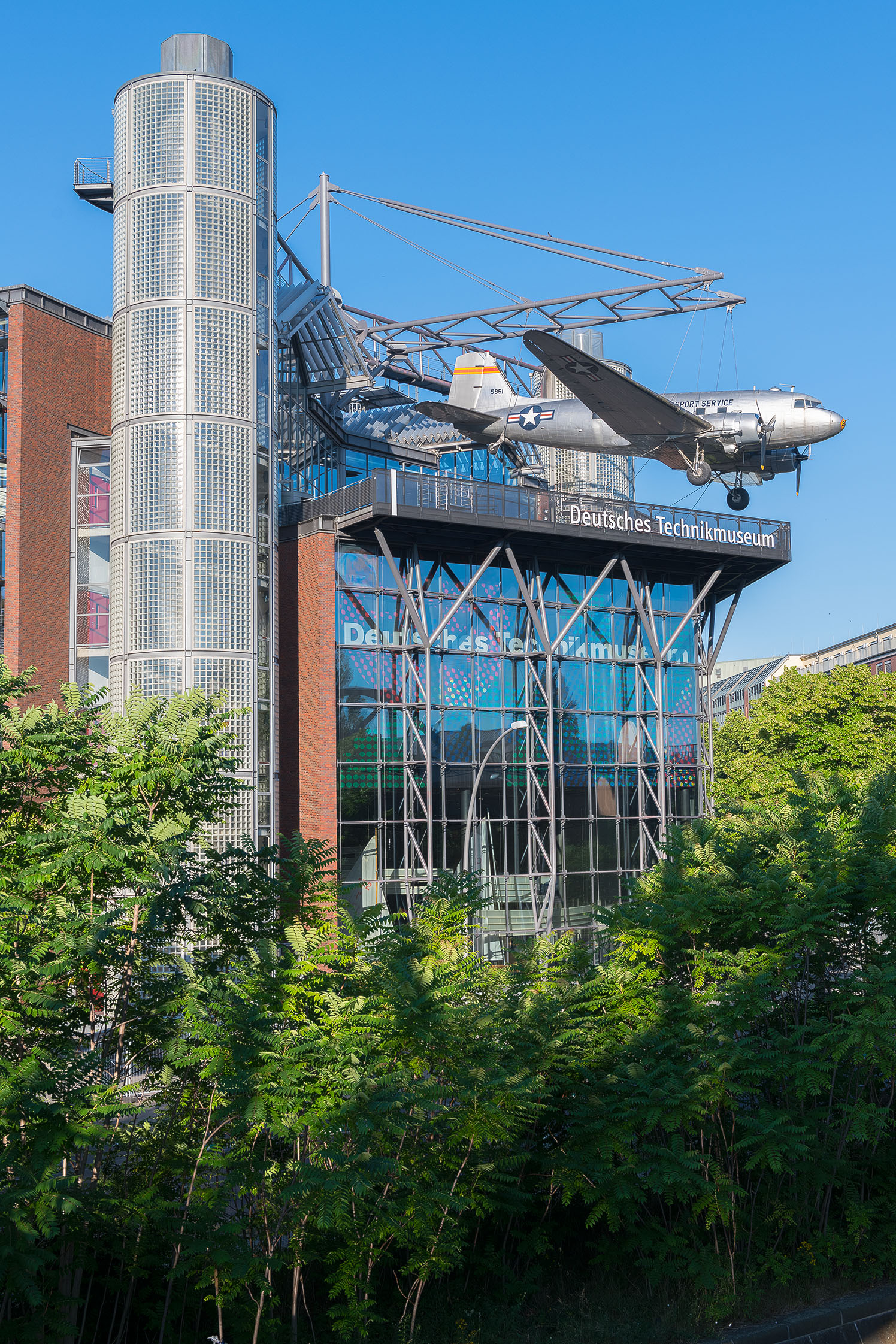 Ein Neubaugebäude mit Glasfassade. An einer Stahlkonstruktion auf dem Dach hängt ein Flugzeug.
