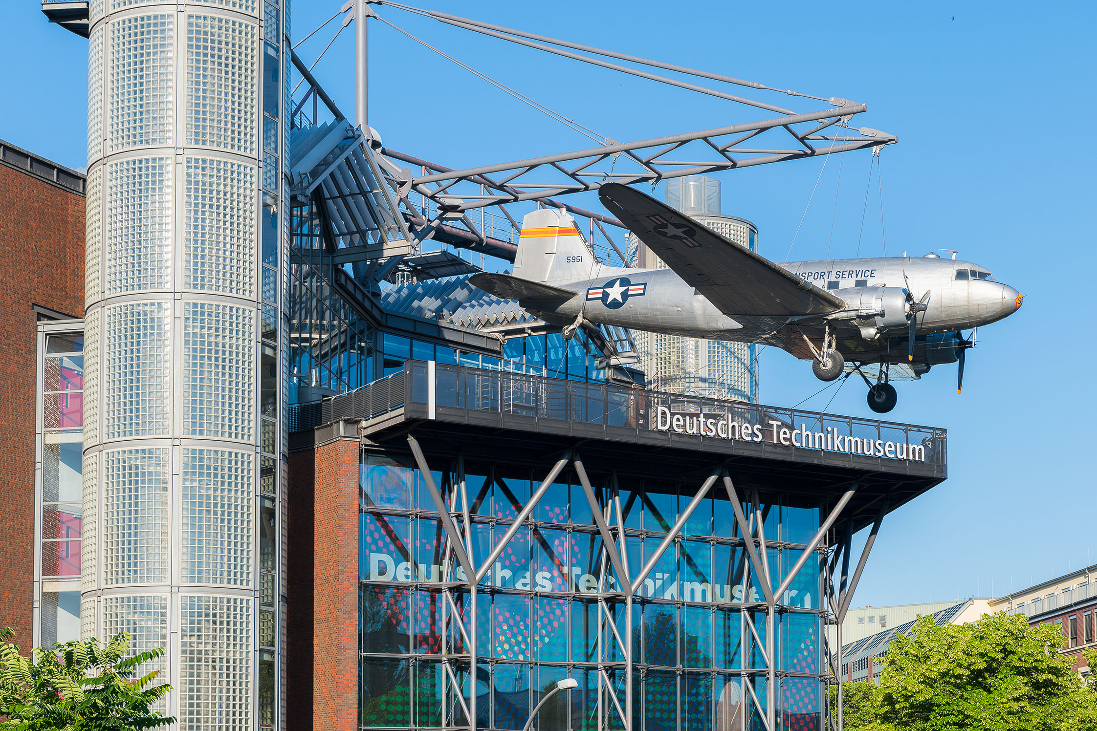 Ein Flugzeug ist mit eines Seilkonstruktion an der Fassade eines verglasten Neubaus angebracht.