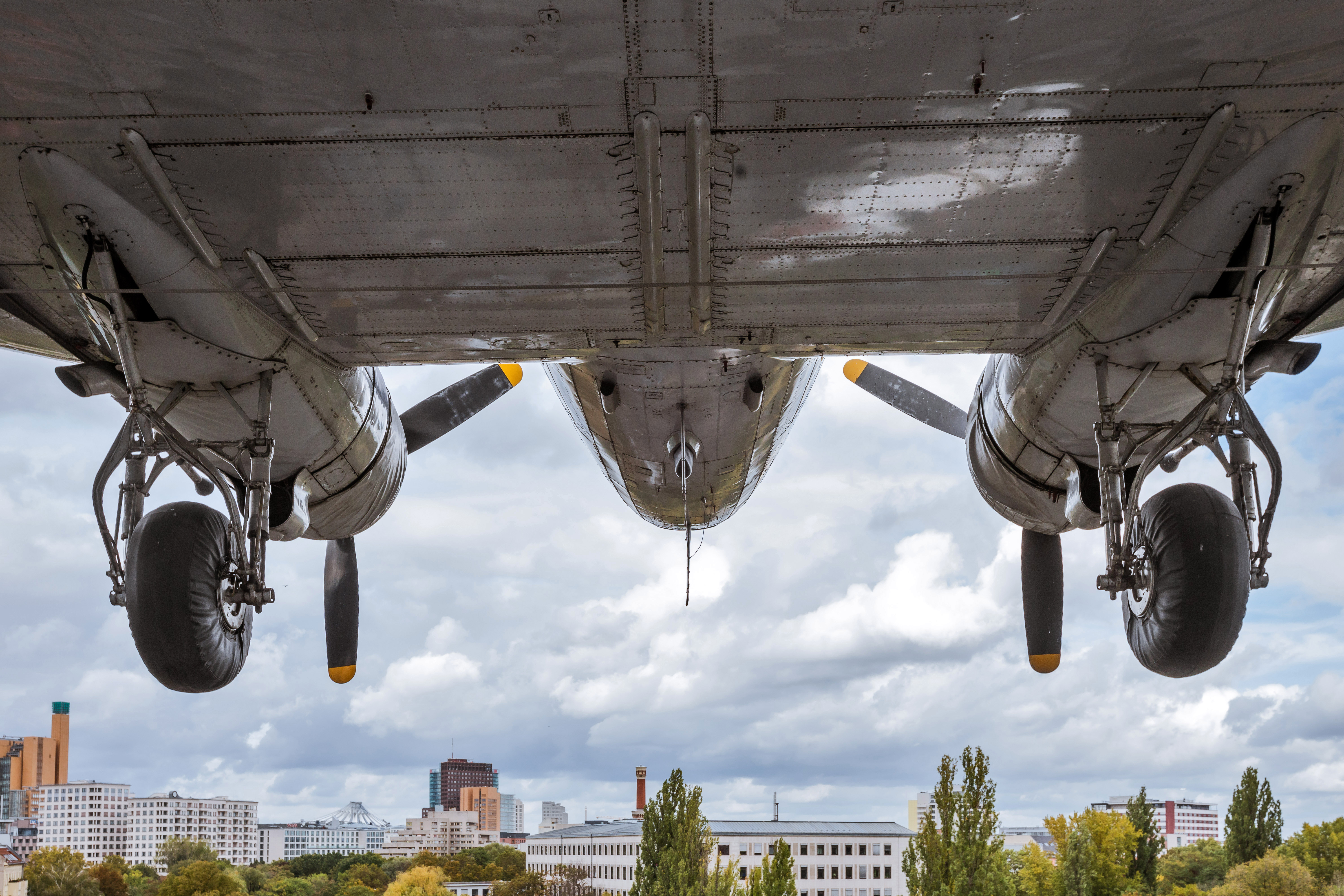 Untersicht auf ein Flugzeug, darunter blickt man auf einen bewölkten Himmel und die Dächer einer Stadt.