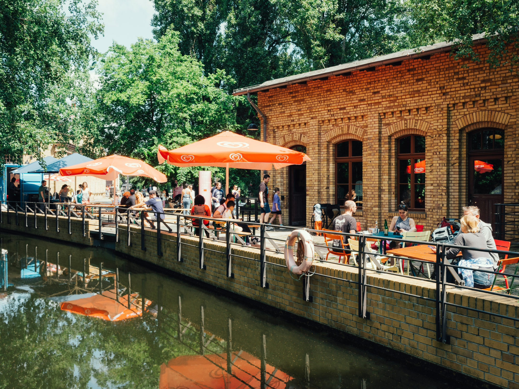A view of the museum pond and a brick building; on the shore, colorful restaurant umbrellas, tables, and benches can be seen, with people sitting at them. In the background is a blue food truck. 