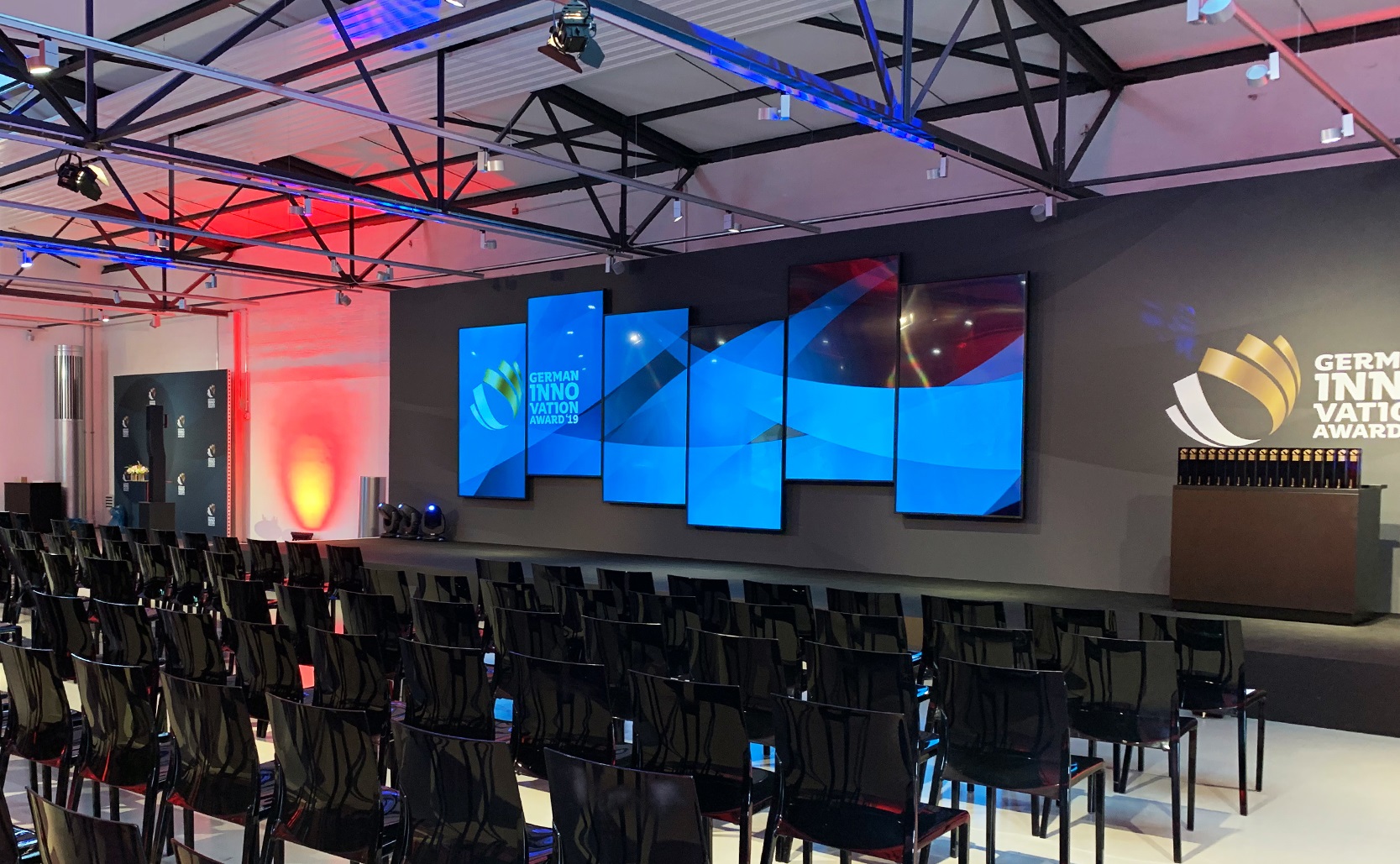 In a hall, rows of chairs and a podium with monitors on the back wall can be seen.